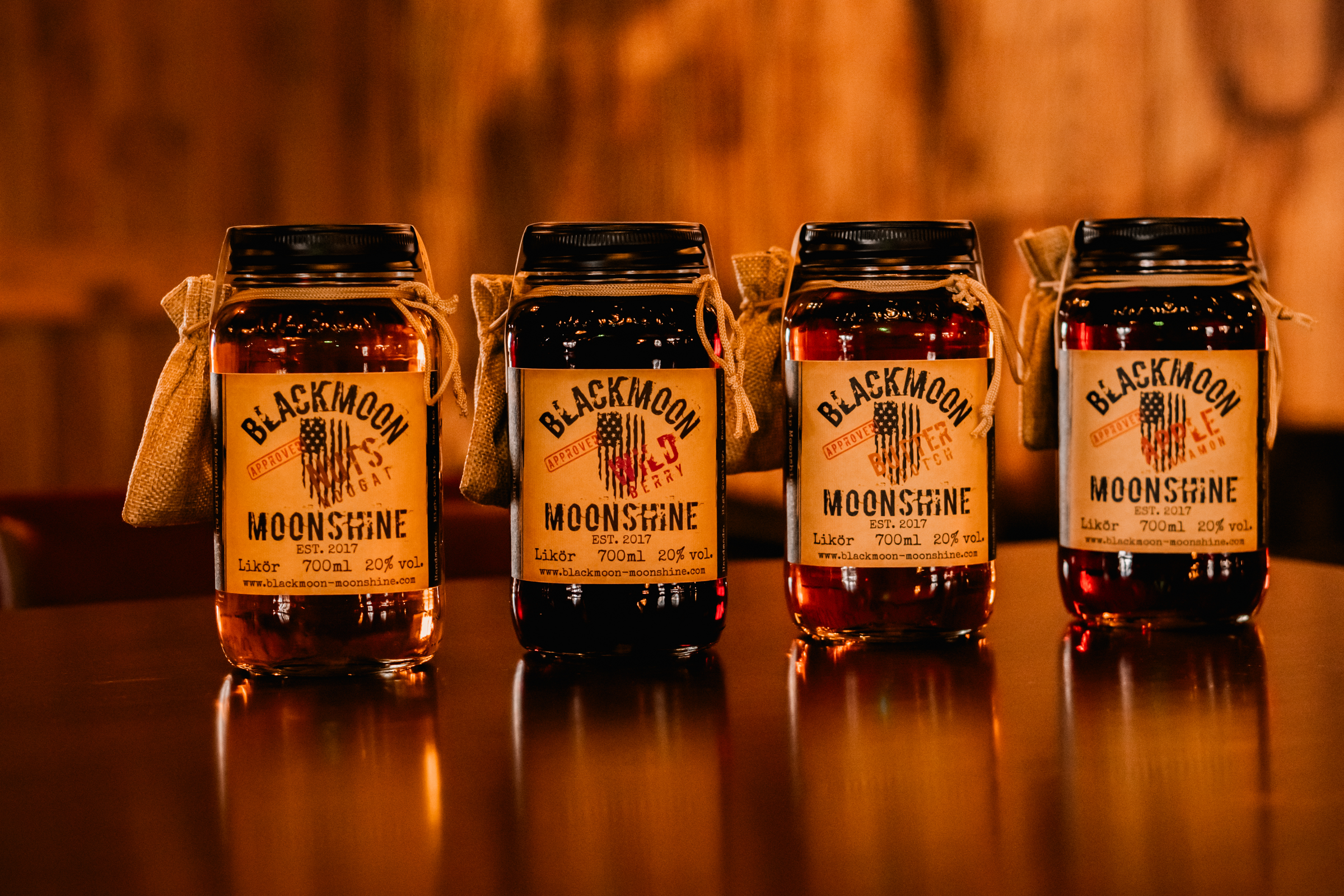 Vier Flaschen BLACKMOON MOONSHINE mit amerikanischer Flagge auf dem Etikett, aufgestellt auf einem Tisch, mit Angaben zu Inhalt (700ml) und Alkoholgehalt (20% vol.).