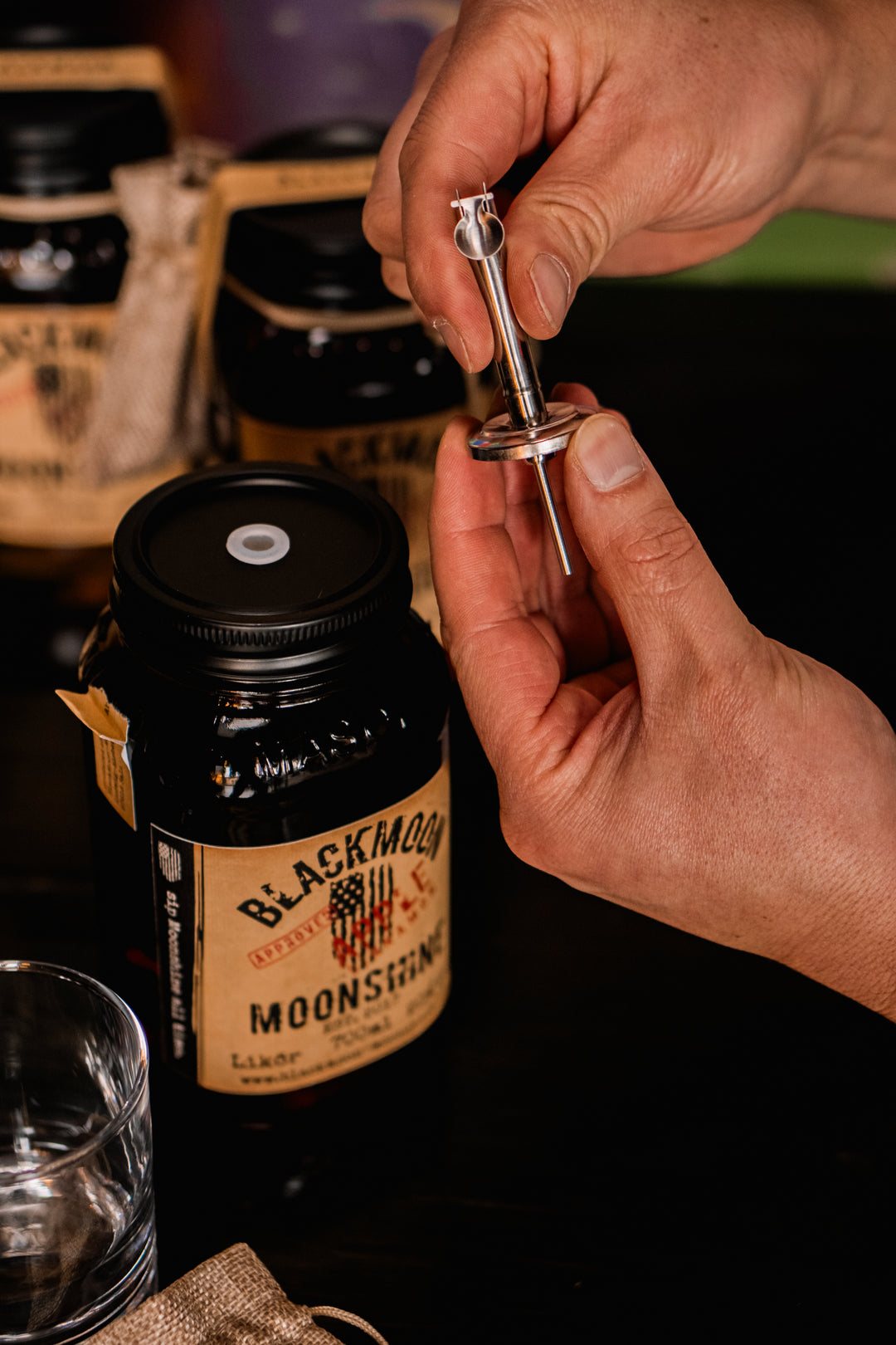 Hand hält einen Metallausgießer über einem BLACKMOON MOONSHINE Jar neben einem leeren Trinkglas.