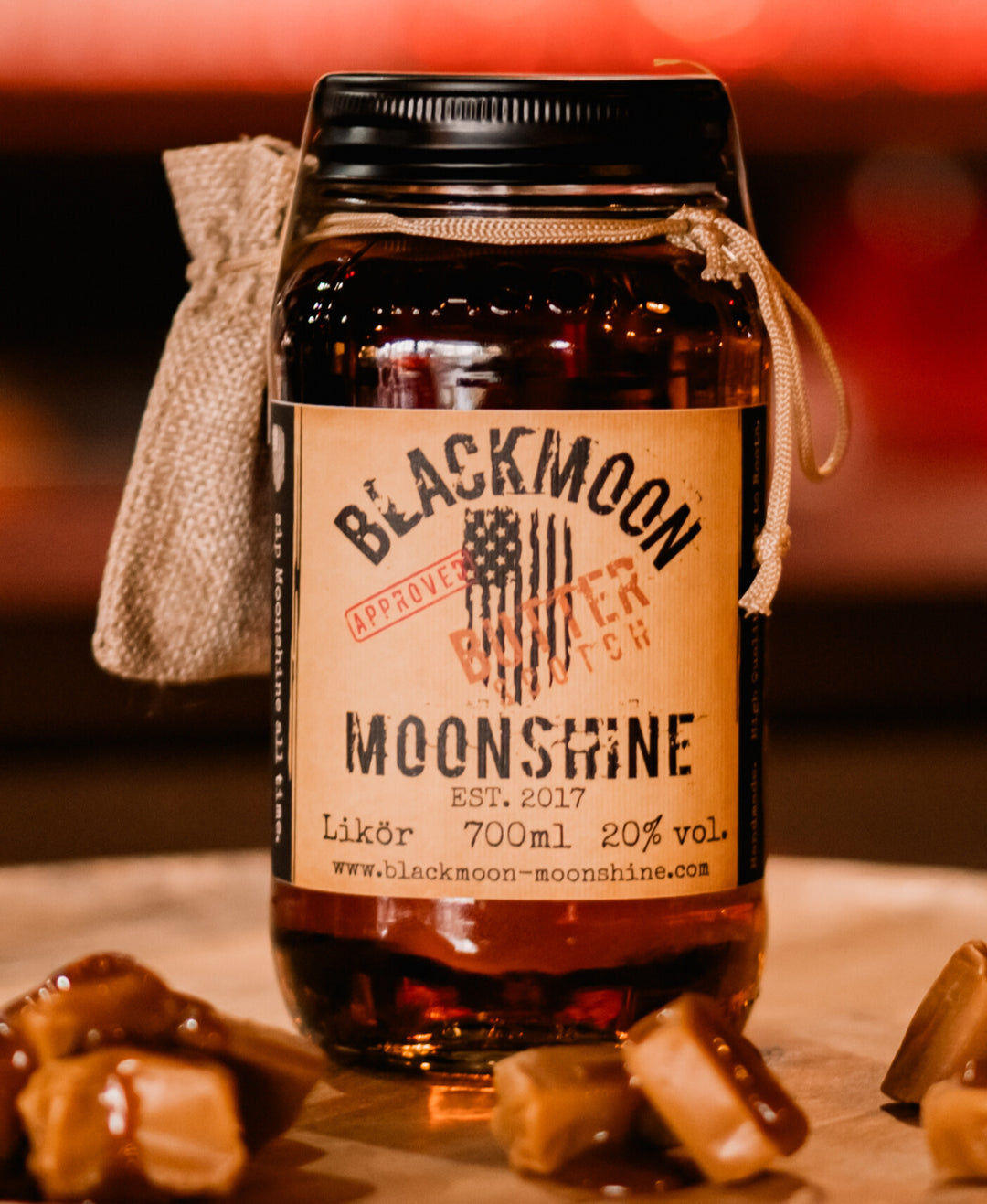 Ein Jar BLACKMOON MOONSHINE mit Karamellbonbons im Vordergrund auf einem Holztisch, Hintergrund unscharf.