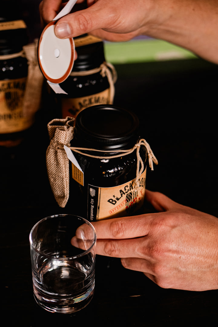 Eine Hand öffnet einen BLACKMOON MOONSHINE Jar neben einem leeren Glas, bereit zum Einschenken.