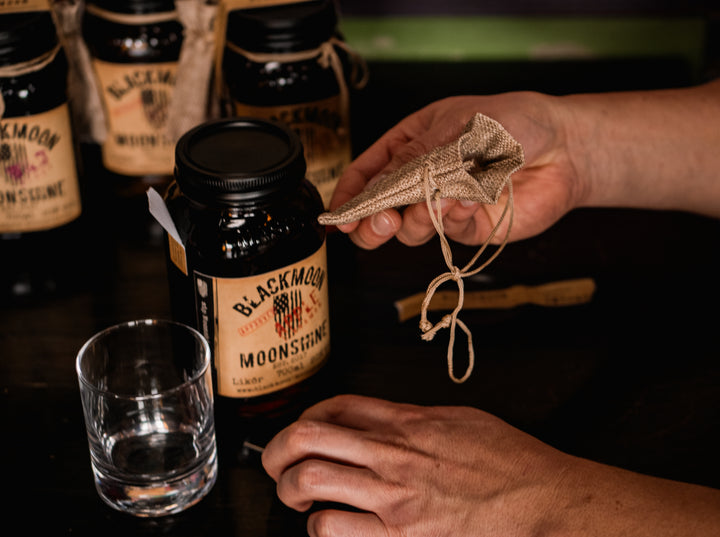 Hände bereiten einen BLACKMOON MOONSHINE Likör vor, mit einem geöffneten Jar und einem leeren Glas auf einem dunklen Holztisch.