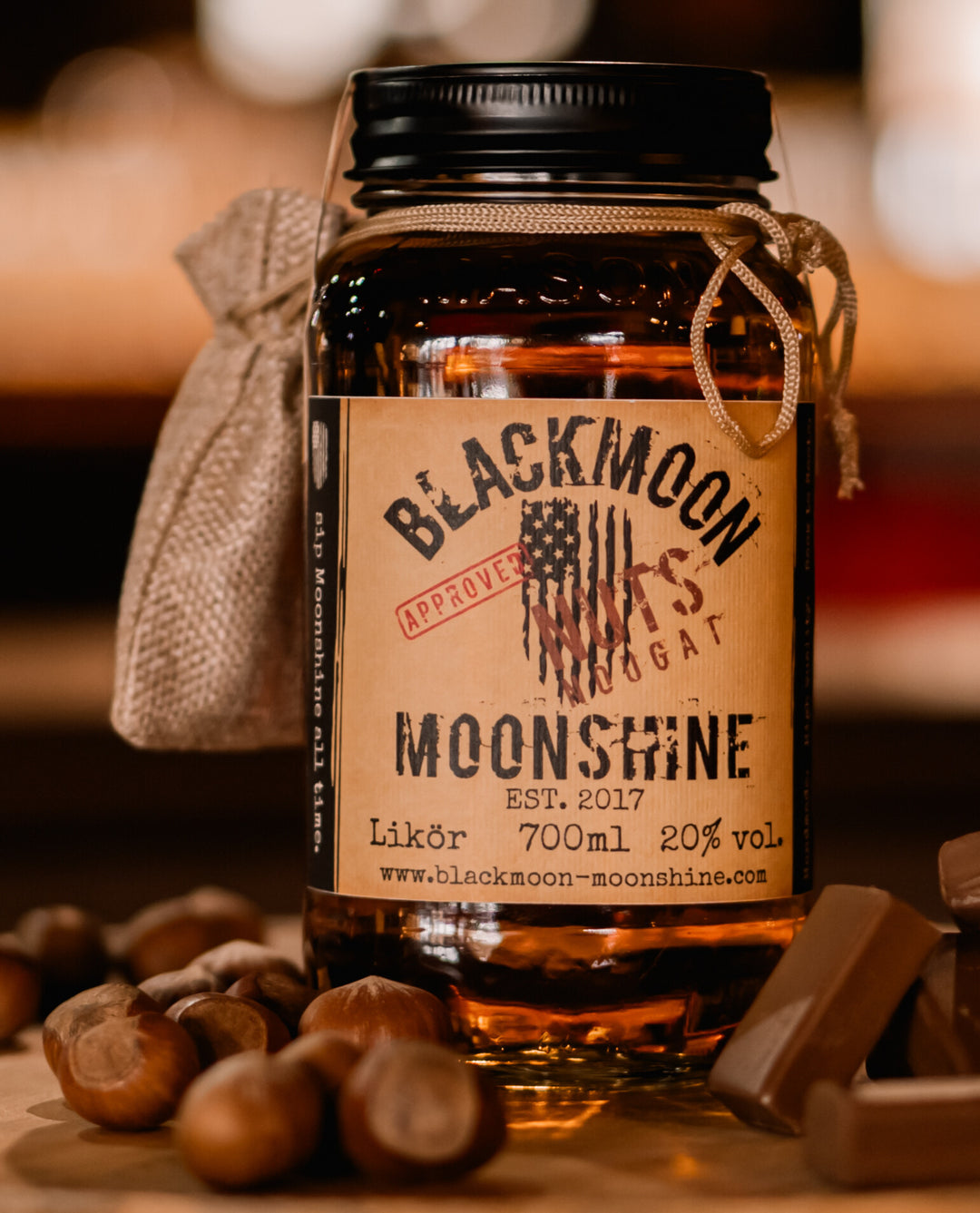 Jar von BLACKMOON NUT NOUGAT MOONSHINE umgeben von Haselnüssen und Karamellstücken auf einem Holztisch.