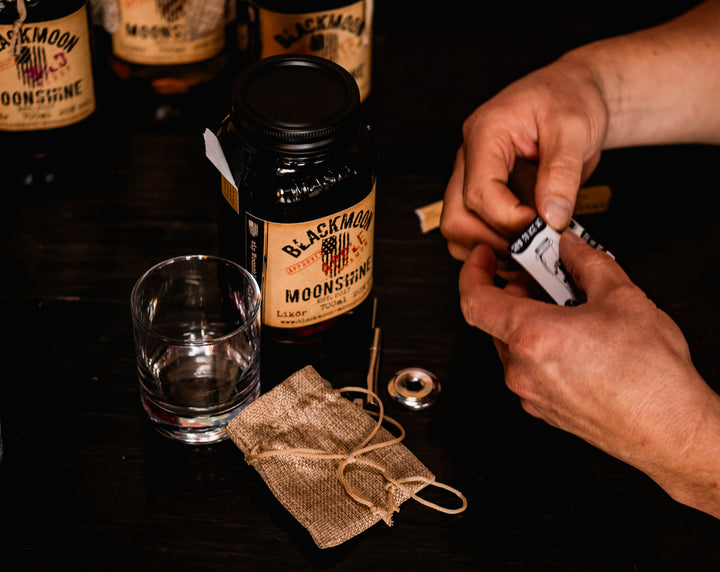 Hände bereiten einen BLACKMOON MOONSHINE Likör vor, mit einem geöffneten Jar und einem leeren Glas auf einem dunklen Holztisch.
