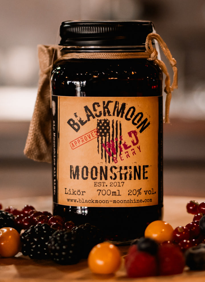 BLACKMOON BERRY MOONSHINE Jar umgeben von frischen Beeren auf einem Holztisch, mit unscharfem Hintergrund.